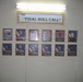 A memorial wall display titled 'The Final Roll Call' is mounted on a light-colored wall, illuminated by a fluorescent light above. The display features twelve framed photographs of military personnel arranged in two rows of six, most photographed against American flag backgrounds. This type of display is a traditional military tribute honoring fallen service members.