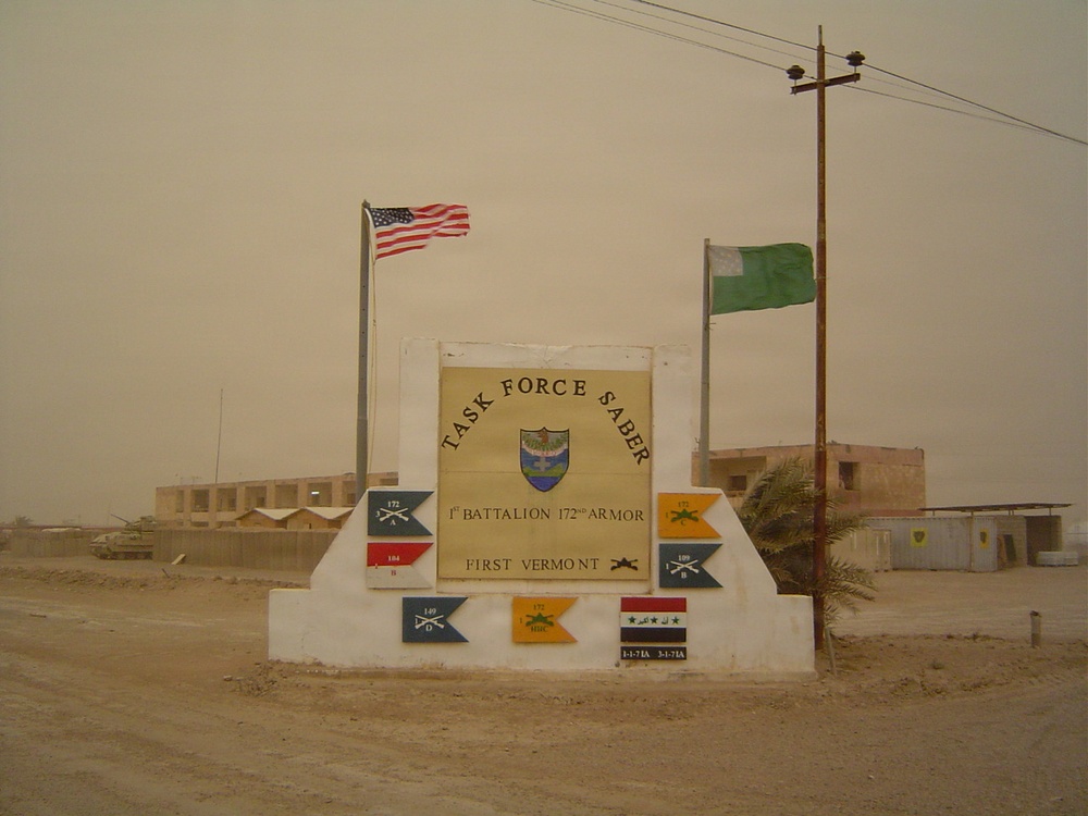 A military installation entrance sign for 'Task Force Saber,' identified as the 1st Battalion, 172nd Armor, First Vermont. The sign is flanked by a US flag and a green flag, with multiple unit guidon pennants displayed on the base. The setting appears to be a desert environment, likely Iraq, with military buildings and a tank visible in the background.