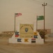 A military installation entrance sign for 'Task Force Saber,' identified as the 1st Battalion, 172nd Armor, First Vermont. The sign is flanked by a US flag and a green flag, with multiple unit guidon pennants displayed on the base. The setting appears to be a desert environment, likely Iraq, with military buildings and a tank visible in the background.