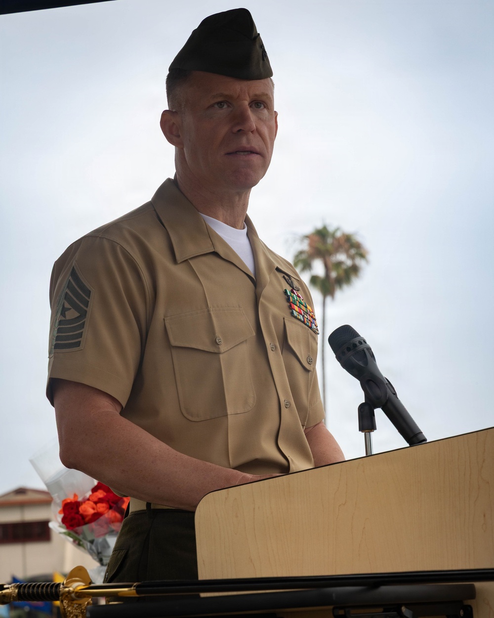 A U.S. Marine Corps senior non-commissioned officer stands at a wooden podium with a microphone, appearing to deliver a speech at an outdoor ceremony. He is wearing the tan USMC Service 'C' uniform with a garrison cover (cover/cap) and displays multiple service ribbons on his chest. A palm tree is visible in the background, suggesting a warm-climate location.