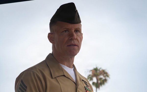A U.S. Marine Corps senior non-commissioned officer stands at a wooden podium with a microphone, appearing to deliver a speech at an outdoor ceremony. He is wearing the tan USMC Service 'C' uniform with a garrison cover (cover/cap) and displays multiple service ribbons on his chest. A palm tree is visible in the background, suggesting a warm-climate location.