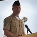 A U.S. Marine Corps senior non-commissioned officer stands at a wooden podium with a microphone, appearing to deliver a speech at an outdoor ceremony. He is wearing the tan USMC Service 'C' uniform with a garrison cover (cover/cap) and displays multiple service ribbons on his chest. A palm tree is visible in the background, suggesting a warm-climate location.