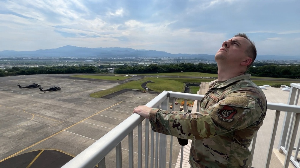 A U.S. Air Force service member in MultiCam/OCP camouflage uniform stands on an elevated observation deck or balcony, looking upward toward the sky. Behind him is a military airfield tarmac with at least two helicopters visible on the ground. Mountains and an urban area are visible in the background, suggesting a location in Asia, possibly Japan.