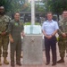 Four military personnel in different uniforms pose together in front of a granite memorial monument with a star emblem and commemorative plaque, located in an outdoor plaza area. The group includes three individuals in U.S. Navy uniforms (two in NWU Type III camouflage and one in an olive green flight suit) and one individual in a light blue dress uniform consistent with an air force service. The setting appears to be a formal military or government compound with trees and manicured landscaping in the background.