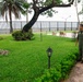 Two military personnel in uniform are standing on a paved walkway, appearing to observe or discuss a memorial monument in a well-maintained outdoor garden area. The setting features lush tropical vegetation, palm trees, and a black iron fence with what appears to be a body of water visible in the background. One individual is wearing a green flight suit and the other is in a light blue service uniform.