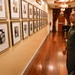 A military officer in a green flight suit with a U.S. flag patch and colonel insignia (eagle rank) is walking through a formal hallway lined with framed portrait photographs on both walls. The hallway appears to be in an official or historic building, featuring hardwood floors, a chandelier, and red curtains at the far end. The officer is pausing to examine the portraits, which appear to be a succession of officials or leaders displayed in chronological order.