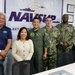 A group of six individuals pose for a formal photo in front of a NAVSUP (Naval Supply Systems Command) Fleet Logistics banner. The group includes two civilians and four military personnel, with three in U.S. Navy uniforms (including one in a flight suit) and one in what appears to be a light blue military uniform. The setting appears to be a conference or administrative office room.