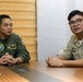 Two military personnel are seated at a conference table engaged in conversation. The individual on the left wears a green flight suit with 'Pacific Partnership' and 'Senior Commander' patches, along with a name tag reading 'Robert Reyes'. The individual on the right wears a tan/khaki military uniform with a name tag reading 'Tero' and multiple service ribbons.