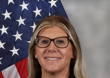 A formal portrait photograph of a woman posing in front of a United States flag against a gray studio background. She is dressed professionally in a black blazer and appears to be smiling directly at the camera. The composition and setting are consistent with an official government or law enforcement headshot.