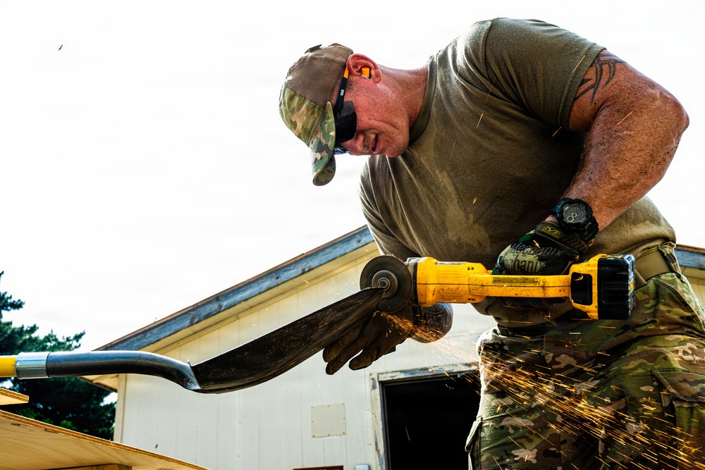 A man in military-style camouflage clothing uses a yellow DeWalt angle grinder to cut through a metal object, producing a shower of bright orange sparks. He is wearing protective gloves (Mechanix brand), sunglasses, a camouflage cap, and has a tribal tattoo visible on his upper arm. The work appears to be taking place outdoors near a building.