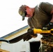 A man in military-style camouflage clothing uses a yellow DeWalt angle grinder to cut through a metal object, producing a shower of bright orange sparks. He is wearing protective gloves (Mechanix brand), sunglasses, a camouflage cap, and has a tribal tattoo visible on his upper arm. The work appears to be taking place outdoors near a building.