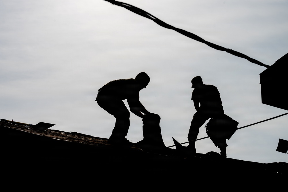 The image shows two workers silhouetted against an overcast sky while working on a rooftop. Both figures are rendered entirely in silhouette due to strong backlighting, making fine details indiscernible. The scene appears to depict roofing or demolition work, with materials visible around the workers.