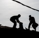 The image shows two workers silhouetted against an overcast sky while working on a rooftop. Both figures are rendered entirely in silhouette due to strong backlighting, making fine details indiscernible. The scene appears to depict roofing or demolition work, with materials visible around the workers.