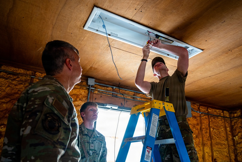 Three military personnel in camouflage uniforms are working inside a wooden-walled structure. One service member is standing on a blue and yellow step ladder installing or wiring a ceiling light fixture, while two others observe from below. The setting appears to be a temporary or expeditionary building under construction or renovation.