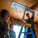 Three military personnel in camouflage uniforms are working inside a wooden-walled structure. One service member is standing on a blue and yellow step ladder installing or wiring a ceiling light fixture, while two others observe from below. The setting appears to be a temporary or expeditionary building under construction or renovation.