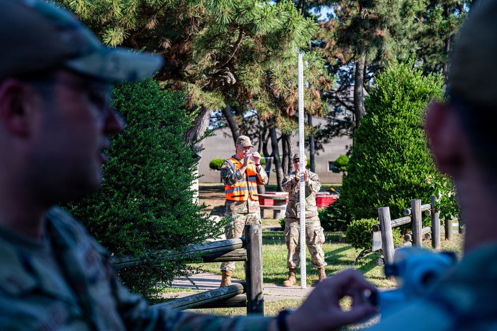 Military personnel in camouflage uniforms are conducting what appears to be a surveying or measurement exercise outdoors. One individual wearing a high-visibility orange safety vest is using binoculars or a sighting device, while another holds a tall graduated surveying rod or staff. The scene takes place in a landscaped area with trees, wooden fencing, and what appears to be a military installation in the background.