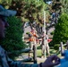 Military personnel in camouflage uniforms are conducting what appears to be a surveying or measurement exercise outdoors. One individual wearing a high-visibility orange safety vest is using binoculars or a sighting device, while another holds a tall graduated surveying rod or staff. The scene takes place in a landscaped area with trees, wooden fencing, and what appears to be a military installation in the background.