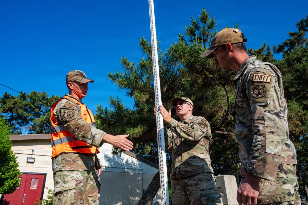 Three military personnel in OCP (Operational Camouflage Pattern) uniforms are working outdoors with a tall measuring rod or surveying staff. One individual wears an orange safety vest, and another has a patch reading 'DIRT' on their uniform. They appear to be conducting a survey or measurement task in a wooded area near a building.