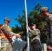 Three military personnel in OCP (Operational Camouflage Pattern) uniforms are working outdoors with a tall measuring rod or surveying staff. One individual wears an orange safety vest, and another has a patch reading 'DIRT' on their uniform. They appear to be conducting a survey or measurement task in a wooded area near a building.