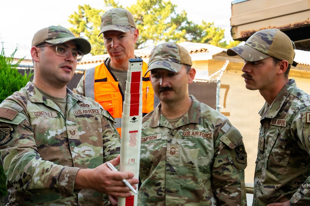 Four individuals are gathered outdoors, with three wearing U.S. Air Force OCP (Operational Camouflage Pattern) uniforms and one wearing a high-visibility orange safety vest. The person on the left, identified by name tape as 'GONZALEZ,' is holding and explaining a measuring rod or survey staff to the others. The individual in the center is identified as 'SIMPSON' with visible Senior Master Sergeant rank insignia.