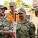 Four individuals are gathered outdoors, with three wearing U.S. Air Force OCP (Operational Camouflage Pattern) uniforms and one wearing a high-visibility orange safety vest. The person on the left, identified by name tape as 'GONZALEZ,' is holding and explaining a measuring rod or survey staff to the others. The individual in the center is identified as 'SIMPSON' with visible Senior Master Sergeant rank insignia.