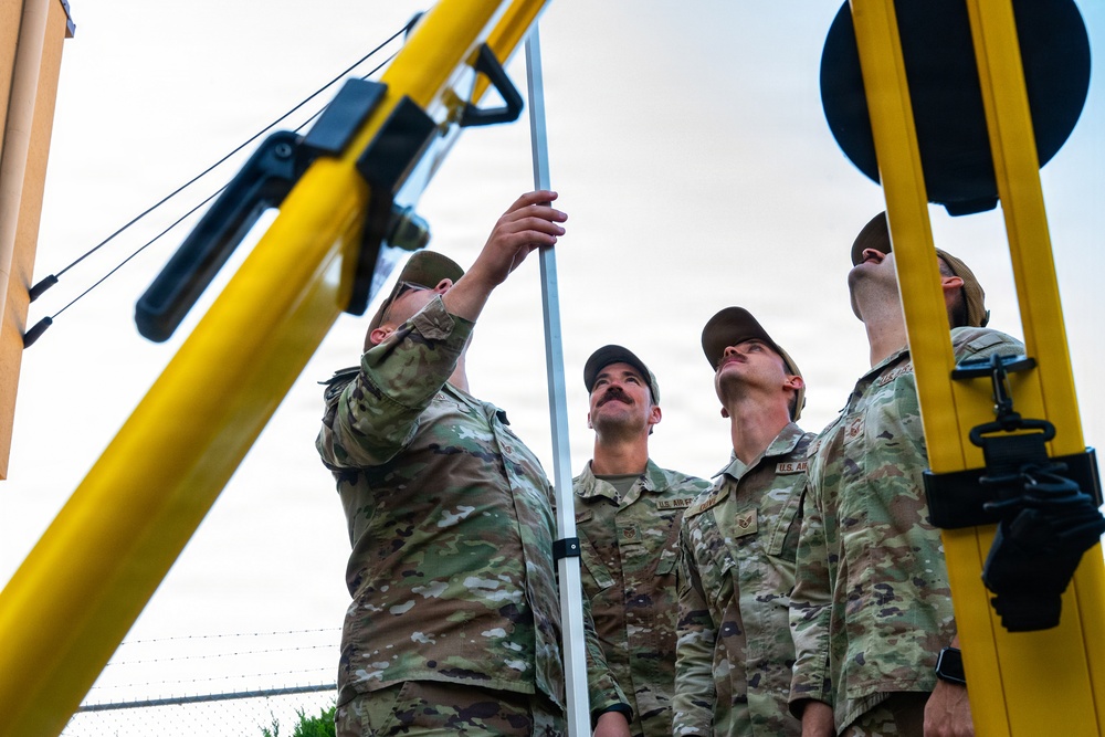 Several U.S. Air Force personnel in OCP (Operational Camouflage Pattern) uniforms are working together to assemble or raise a metal pole or mast structure. They are positioned around yellow-painted equipment, likely a portable lifting or rigging apparatus. The image is taken from a low angle, looking upward, emphasizing the height of the structure being erected.