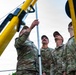 Several U.S. Air Force personnel in OCP (Operational Camouflage Pattern) uniforms are working together to assemble or raise a metal pole or mast structure. They are positioned around yellow-painted equipment, likely a portable lifting or rigging apparatus. The image is taken from a low angle, looking upward, emphasizing the height of the structure being erected.