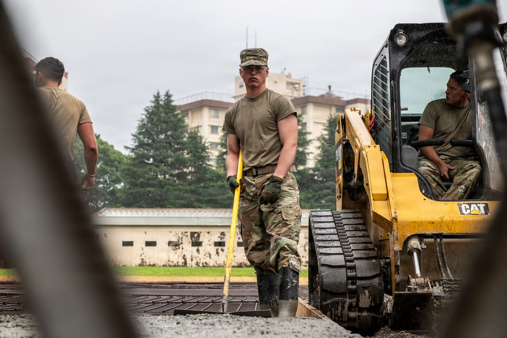 Military personnel in OCP (Operational Camouflage Pattern) uniforms are conducting construction or engineering work at what appears to be a military installation. A central figure is using a long-handled tool, likely a rake or screed, to level gravel or aggregate material, while another service member operates a yellow CAT skid steer loader nearby. The setting includes buildings and trees consistent with a military base environment.