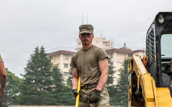 Military personnel in OCP (Operational Camouflage Pattern) uniforms are conducting construction or engineering work at what appears to be a military installation. A central figure is using a long-handled tool, likely a rake or screed, to level gravel or aggregate material, while another service member operates a yellow CAT skid steer loader nearby. The setting includes buildings and trees consistent with a military base environment.