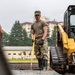 Military personnel in OCP (Operational Camouflage Pattern) uniforms are conducting construction or engineering work at what appears to be a military installation. A central figure is using a long-handled tool, likely a rake or screed, to level gravel or aggregate material, while another service member operates a yellow CAT skid steer loader nearby. The setting includes buildings and trees consistent with a military base environment.