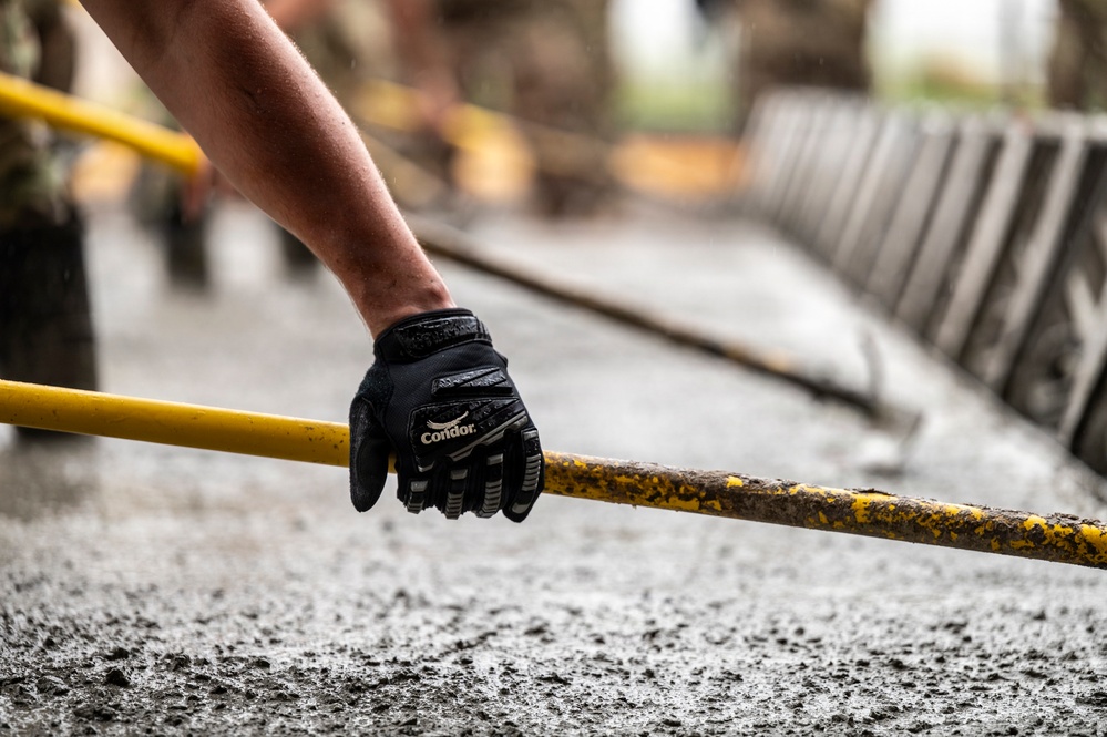 A person wearing a black Condor tactical/work glove grips a yellow metal railing or bar, likely at a construction or industrial site. The surface below appears to be freshly poured or wet concrete. The background shows additional yellow railings and metal structures in a blurred depth-of-field composition.