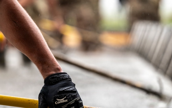 A person wearing a black Condor tactical/work glove grips a yellow metal railing or bar, likely at a construction or industrial site. The surface below appears to be freshly poured or wet concrete. The background shows additional yellow railings and metal structures in a blurred depth-of-field composition.
