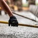 A person wearing a black Condor tactical/work glove grips a yellow metal railing or bar, likely at a construction or industrial site. The surface below appears to be freshly poured or wet concrete. The background shows additional yellow railings and metal structures in a blurred depth-of-field composition.