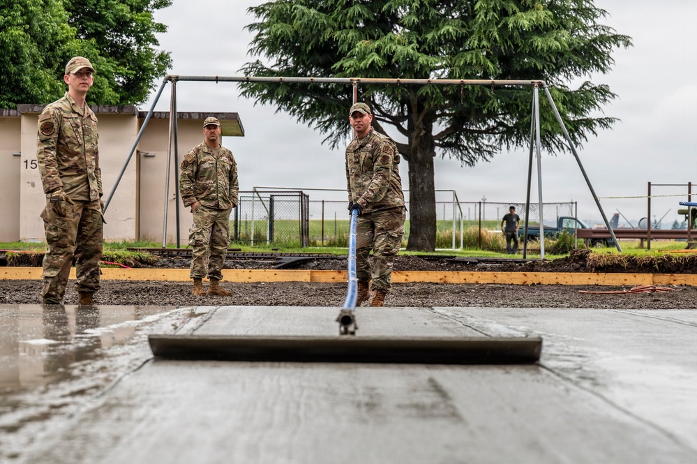 Three military personnel in MultiCam/OCP camouflage uniforms are engaged in a concrete pouring and finishing operation outdoors. One service member is actively using a concrete screed/float tool to level freshly poured concrete, while two others observe. The setting appears to be a military installation, with a swing set frame, chain-link fencing, and what appears to be an airfield visible in the background.