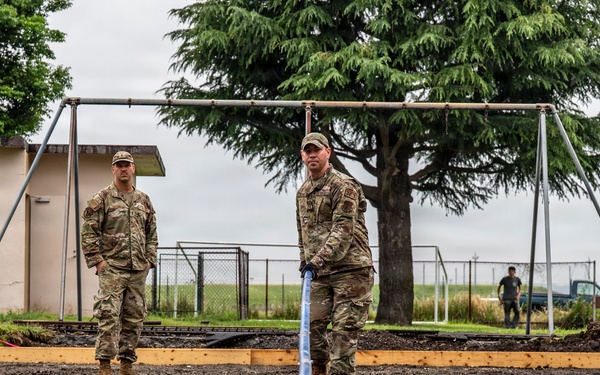 Three military personnel in MultiCam/OCP camouflage uniforms are engaged in a concrete pouring and finishing operation outdoors. One service member is actively using a concrete screed/float tool to level freshly poured concrete, while two others observe. The setting appears to be a military installation, with a swing set frame, chain-link fencing, and what appears to be an airfield visible in the background.
