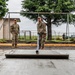 Three military personnel in MultiCam/OCP camouflage uniforms are engaged in a concrete pouring and finishing operation outdoors. One service member is actively using a concrete screed/float tool to level freshly poured concrete, while two others observe. The setting appears to be a military installation, with a swing set frame, chain-link fencing, and what appears to be an airfield visible in the background.