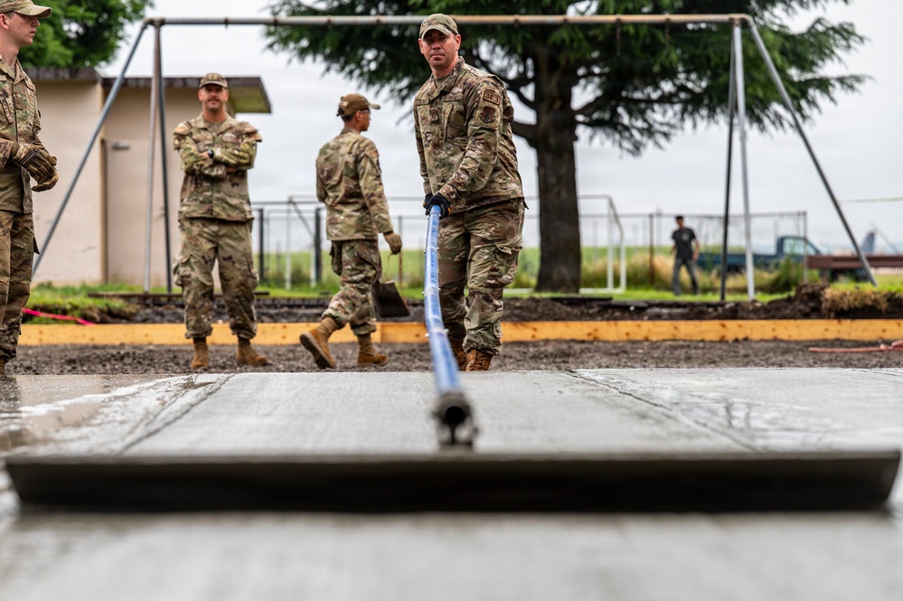 Military personnel in OCP (Operational Camouflage Pattern) uniforms are performing concrete work, likely on a base or installation. One service member in the foreground is using a screed tool to level freshly poured concrete, while others observe or assist in the background. The setting appears to be a construction or renovation project near a playground area.