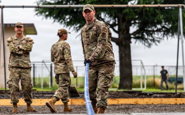 Military personnel in OCP (Operational Camouflage Pattern) uniforms are performing concrete work, likely on a base or installation. One service member in the foreground is using a screed tool to level freshly poured concrete, while others observe or assist in the background. The setting appears to be a construction or renovation project near a playground area.