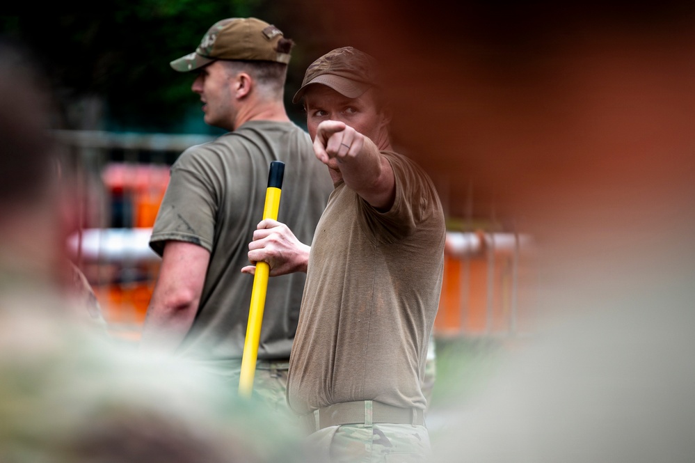Two individuals wearing olive/tan military-style clothing are visible in an outdoor setting. The foreground figure is pointing with one hand while holding a yellow-handled tool or implement, appearing to give directions or instructions. The background figure wears a military-style cap and stands nearby.