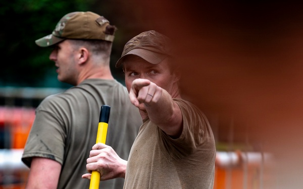 Two individuals wearing olive/tan military-style clothing are visible in an outdoor setting. The foreground figure is pointing with one hand while holding a yellow-handled tool or implement, appearing to give directions or instructions. The background figure wears a military-style cap and stands nearby.