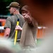 Two individuals wearing olive/tan military-style clothing are visible in an outdoor setting. The foreground figure is pointing with one hand while holding a yellow-handled tool or implement, appearing to give directions or instructions. The background figure wears a military-style cap and stands nearby.