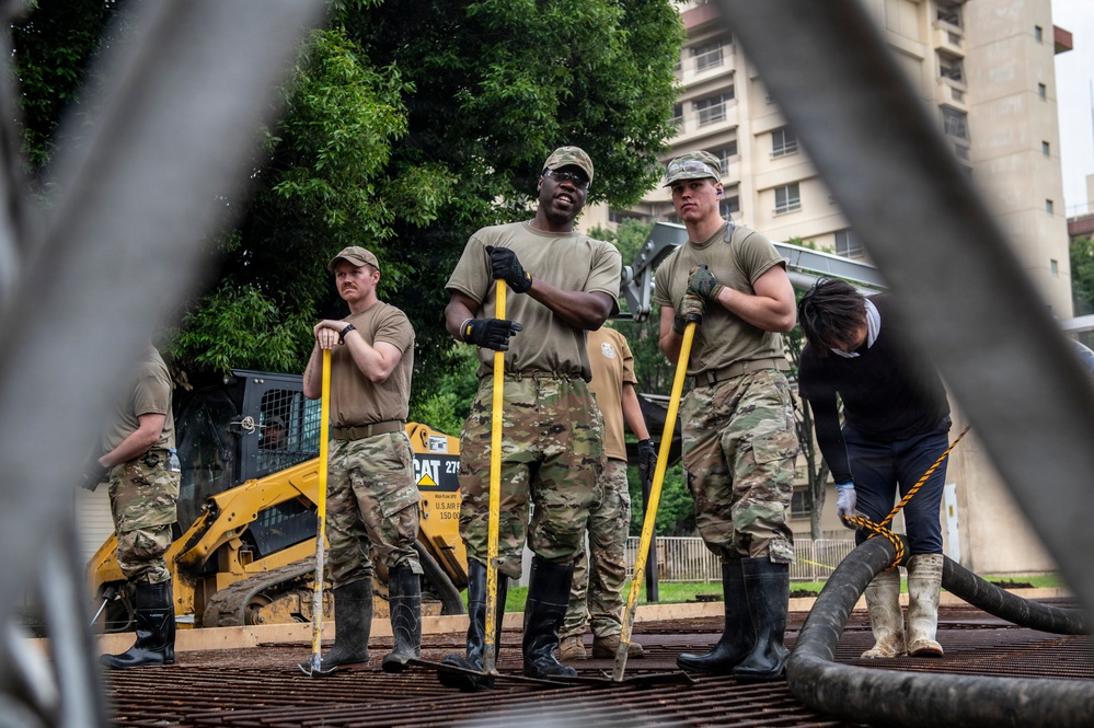 Military personnel in U.S. Army camouflage uniforms are engaged in what appears to be a flood response or construction/cleanup operation. They are standing on a metal grate surface holding long-handled tools, wearing rubber boots and work gloves, with a Caterpillar skid-steer loader visible in the background. The image is photographed through a chain-link fence, suggesting a secured perimeter around the work area.