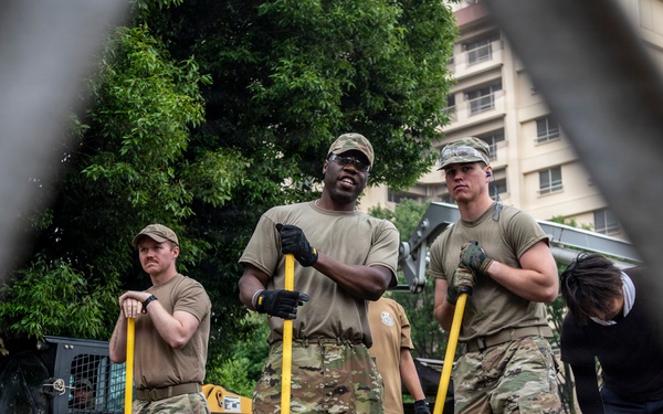 Military personnel in U.S. Army camouflage uniforms are engaged in what appears to be a flood response or construction/cleanup operation. They are standing on a metal grate surface holding long-handled tools, wearing rubber boots and work gloves, with a Caterpillar skid-steer loader visible in the background. The image is photographed through a chain-link fence, suggesting a secured perimeter around the work area.