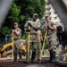 Military personnel in U.S. Army camouflage uniforms are engaged in what appears to be a flood response or construction/cleanup operation. They are standing on a metal grate surface holding long-handled tools, wearing rubber boots and work gloves, with a Caterpillar skid-steer loader visible in the background. The image is photographed through a chain-link fence, suggesting a secured perimeter around the work area.