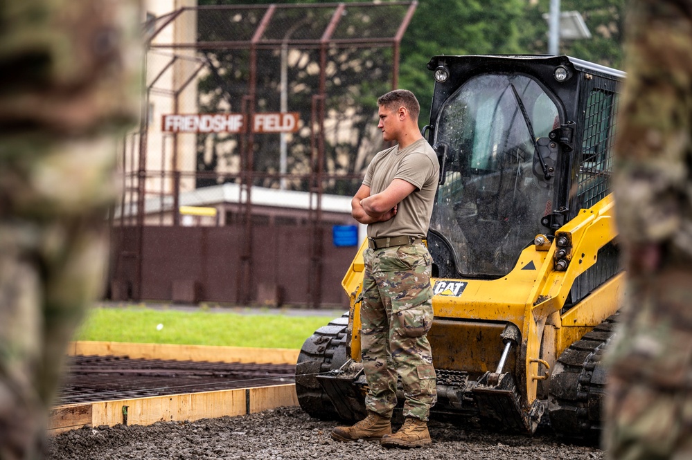 A military service member in OCP (Operational Camouflage Pattern) uniform stands with arms crossed beside a yellow CAT (Caterpillar) compact track loader at a construction site. The work appears to be taking place near a location identified as 'Friendship Field,' visible on a sign in the background. The soldier appears to be overseeing or pausing during construction or grading work.