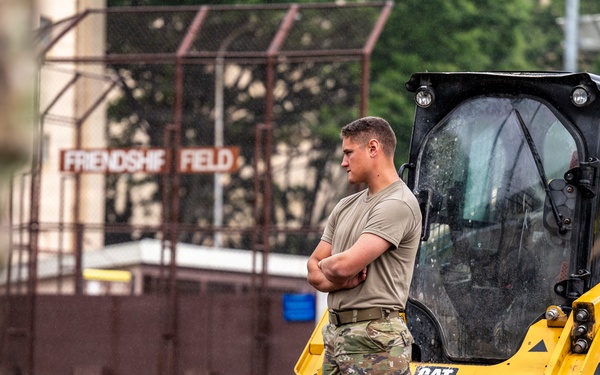 A military service member in OCP (Operational Camouflage Pattern) uniform stands with arms crossed beside a yellow CAT (Caterpillar) compact track loader at a construction site. The work appears to be taking place near a location identified as 'Friendship Field,' visible on a sign in the background. The soldier appears to be overseeing or pausing during construction or grading work.
