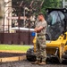 A military service member in OCP (Operational Camouflage Pattern) uniform stands with arms crossed beside a yellow CAT (Caterpillar) compact track loader at a construction site. The work appears to be taking place near a location identified as 'Friendship Field,' visible on a sign in the background. The soldier appears to be overseeing or pausing during construction or grading work.
