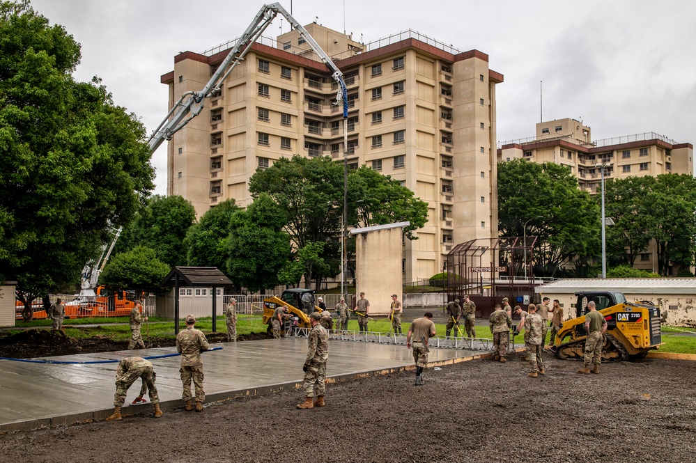 Military personnel in camouflage uniforms are engaged in a concrete pouring and finishing operation on what appears to be a military installation. A concrete pump boom extends over the work area, and a CAT 279D skid steer loader is visible on the right side. The work is taking place in front of a large multi-story beige building with a sign reading 'Friendship Field' visible in the background.