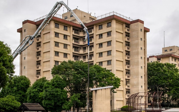 Military personnel in camouflage uniforms are engaged in a concrete pouring and finishing operation on what appears to be a military installation. A concrete pump boom extends over the work area, and a CAT 279D skid steer loader is visible on the right side. The work is taking place in front of a large multi-story beige building with a sign reading 'Friendship Field' visible in the background.