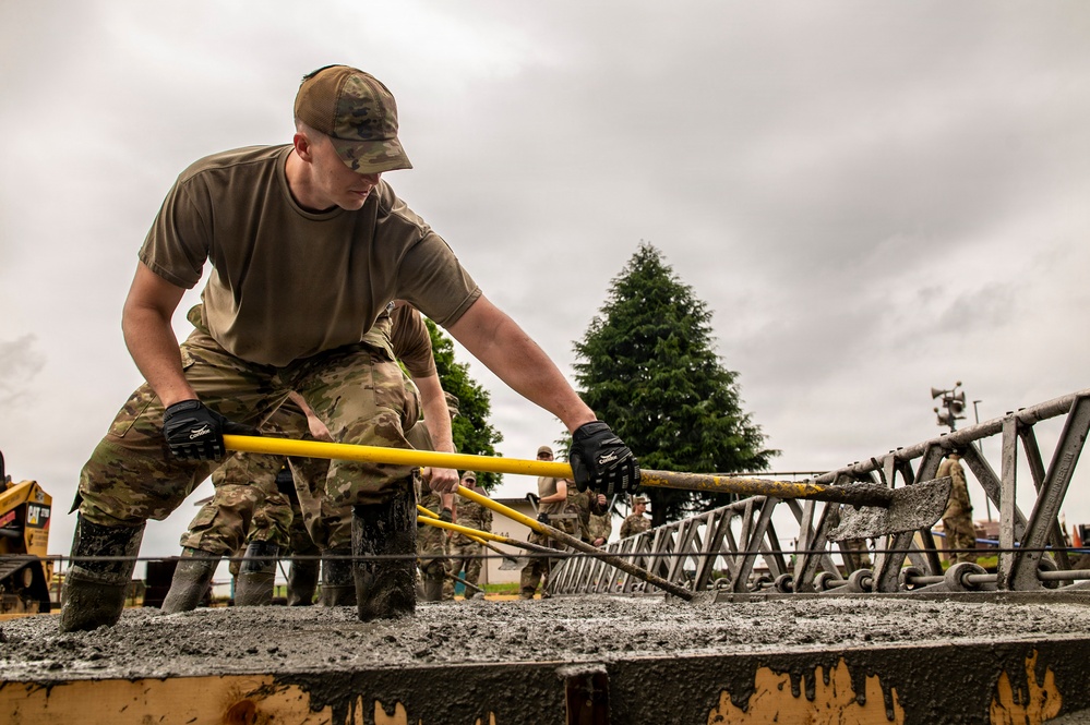 Military personnel in MultiCam/OCP camouflage uniforms are working to level or screed freshly poured concrete using a yellow screed bar. The foreground soldier is leaning forward with both gloved hands gripping the tool, actively smoothing the wet concrete surface. A metal rebar/reinforcement framework is visible embedded in the concrete, and additional personnel and construction equipment are visible in the background.